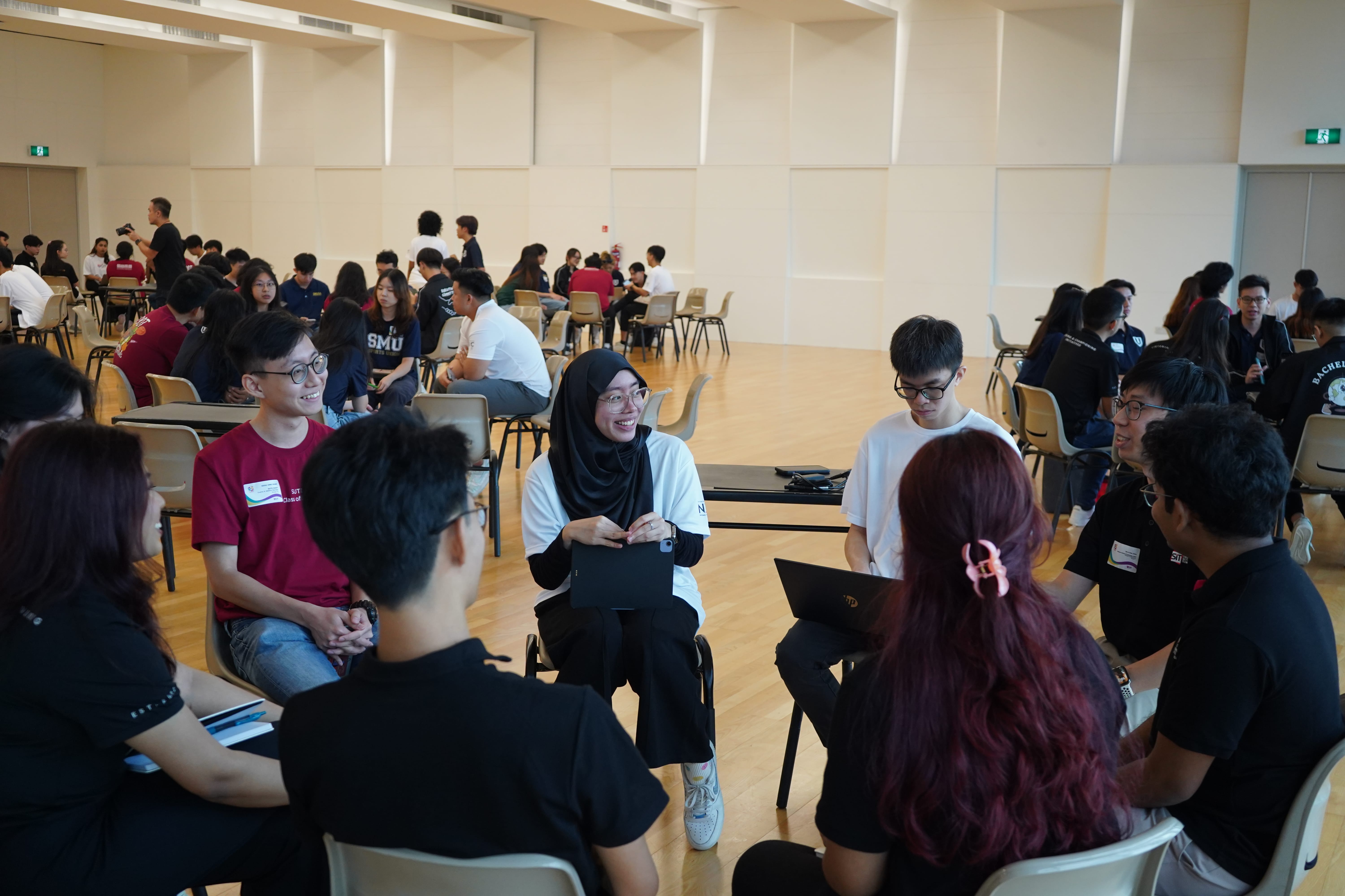 Large room with groups of students sitting in circles, some using laptops, wearing SMU shirts.